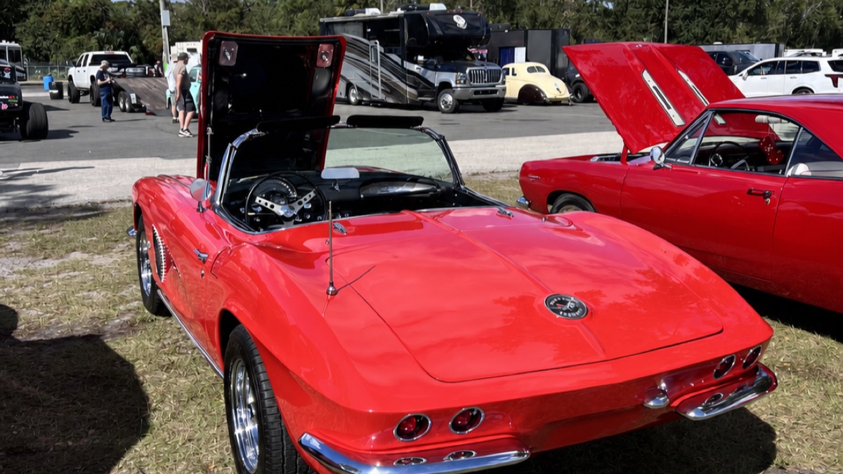 A red early-1960s Corvette convertible with its hood up at a sunny outdoor car show