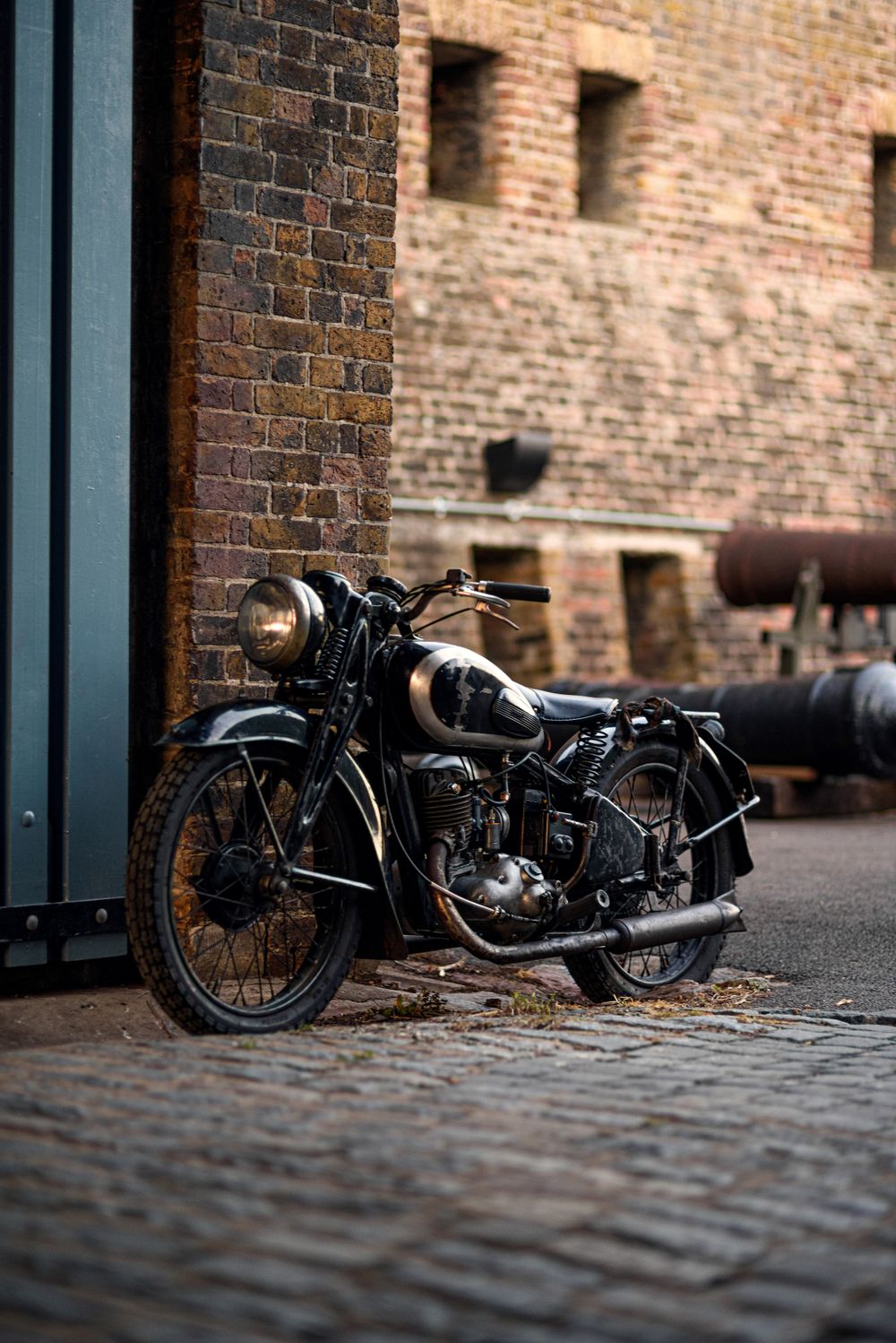 A vintage black motorcycle with a brass headlamp parked against a weathered brick wall