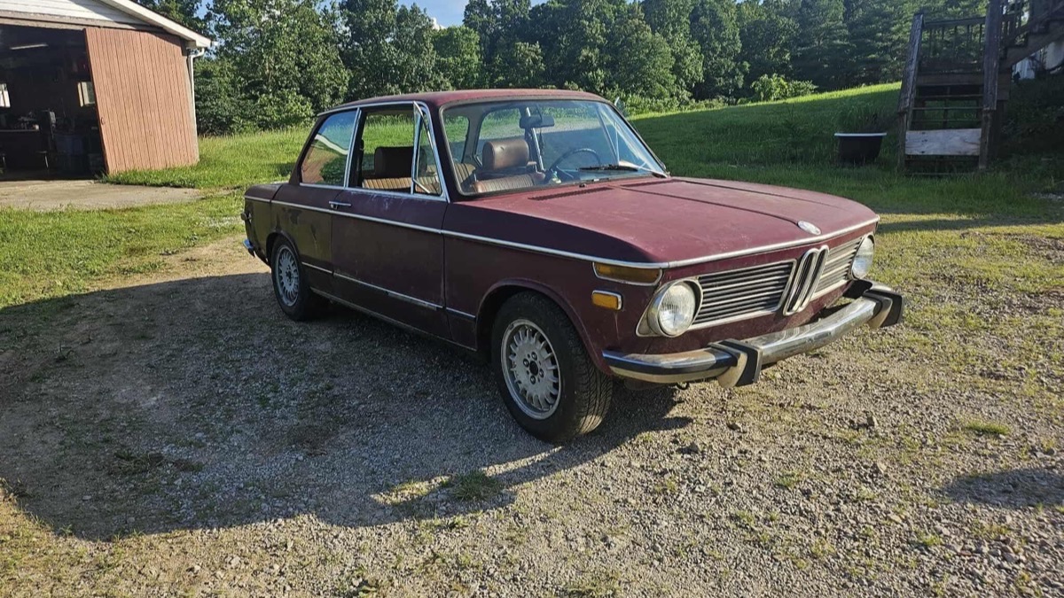 A maroon 1970s BMW 2002 parked on gravel beside a barn, green hills behind it