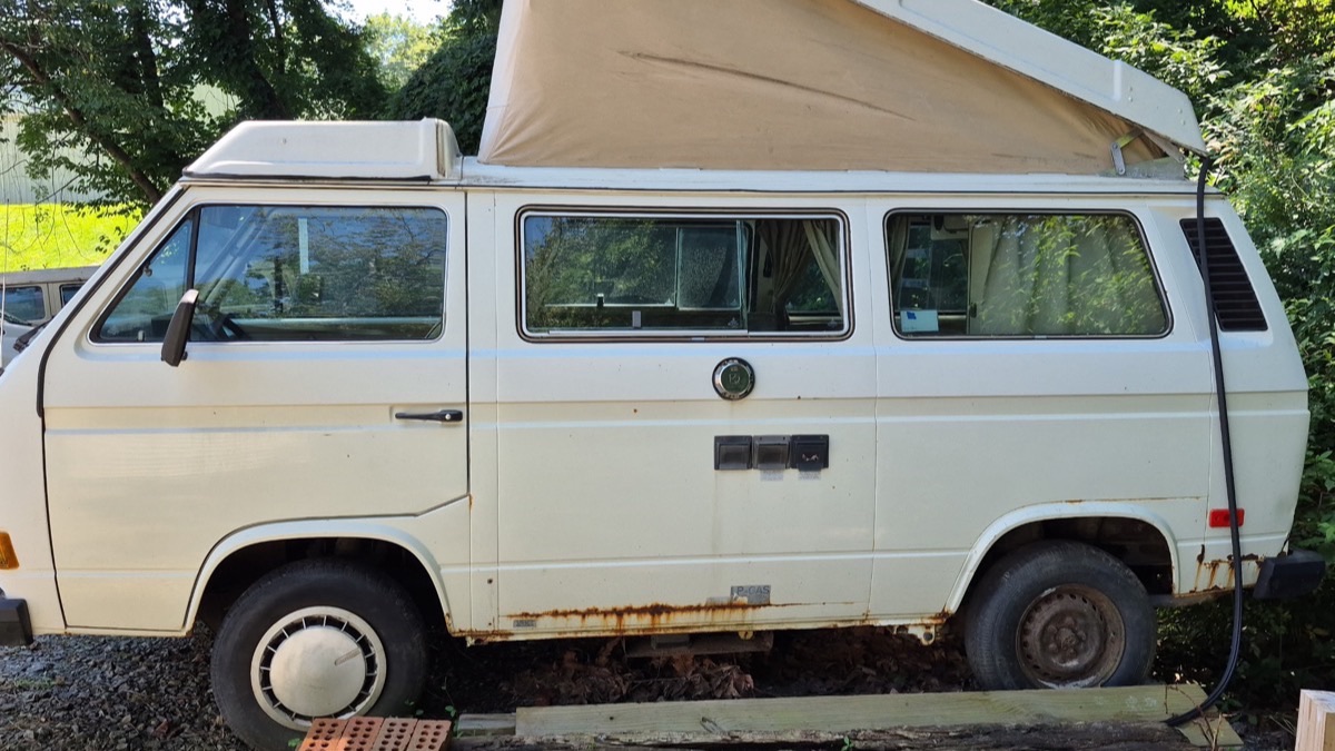 A white Volkswagen Vanagon camper with its pop-top raised, sitting on blocks in an overgrown yard